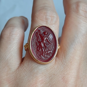 Puede incluir: Un anillo de oro con un camafeo rojo que representa a San Miguel Arcángel matando a un dragón. El camafeo está engastado en un bisel ovalado de oro.
