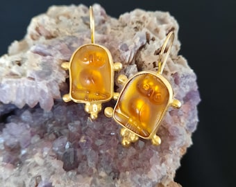 Puede incluir: Un par de pendientes de oro con colgantes de búho de ámbar. Los búhos están tallados con detalle y tienen un color cálido, similar al de la miel.