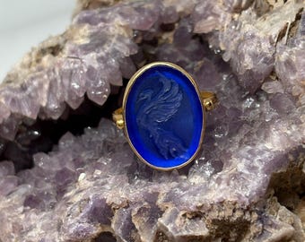May include: A gold ring with an oval, deep blue stone featuring a carved bird design. The ring is set against a backdrop of a rough, purple-hued rock formation, creating a striking contrast of colors and textures. The ring's design is classic.