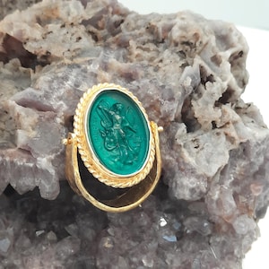 May include: A gold ring with a green cameo featuring a depiction of an angel with the word "MICHAEL" inscribed around the cameo.