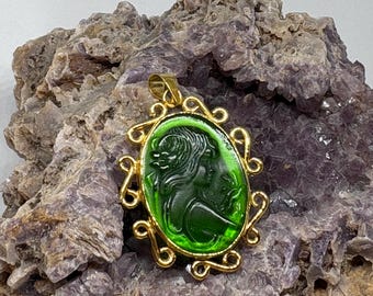 Puede incluir: Colgante dorado con un camafeo verde ovalado que representa el perfil de una mujer. El camafeo está engastado en un marco dorado decorativo. El colgante se muestra sobre un fondo texturizado de color púrpura-gris.