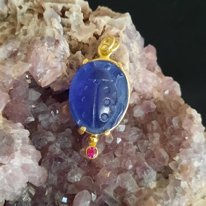 May include: A gold pendant featuring a blue glass ladybug with a red gemstone accent. The pendant is displayed on a bed of pink crystals.