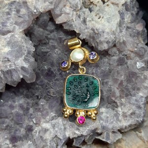 May include: Gold pendant featuring a green cameo with a peacock design, surrounded by small purple and pink gemstones.