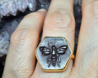 May include: A gold ring with a hexagonal bezel featuring a dark gray cameo of a bee. The bee is detailed with wings, legs, and a stinger.