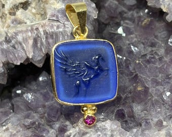 May include: A gold-colored pendant with a square, deep blue center featuring a carved bird design. A small, round, pink gemstone hangs below the square. The pendant is set against a purple, crystalline background.