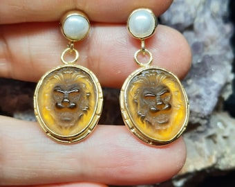 May include: A pair of gold earrings with a carved lion head design in amber glass and a white pearl accent.