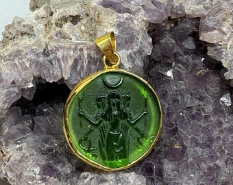 Puede incluir: Colgante dorado con un inserto de vidrio verde circular. El inserto presenta una figura tallada con múltiples brazos, una luna creciente y antorchas. El colgante está sobre un fondo de cristal púrpura.