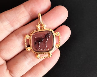 Puede incluir: Un colgante de oro con un camafeo de forma cuadrada que presenta una representación de un toro en vidrio morado. El camafeo está rodeado por un marco de oro con un diseño de cuerda.