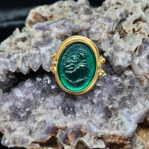 Puede incluir: Un anillo de oro con una piedra ovalada de color verde esmeralda. La piedra presenta una imagen tallada de una figura religiosa, con la inscripción "ST ANTHONY" visible. El anillo está colocado sobre un fondo de formación cristalina morada y blanca.