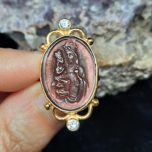 Puede incluir: Anillo de oro con un camafeo rojo que representa a la Virgen María y al niño Jesús. El camafeo está rodeado por un diseño de filigrana dorada y adornado con dos piedras de circonita cúbica transparentes.