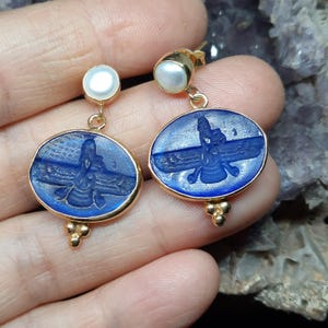 May include: A pair of gold earrings with blue glass inlays featuring a winged creature design. Each earring has a small white pearl at the top.