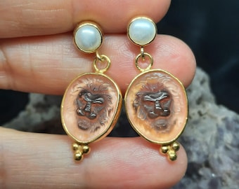 May include: A pair of gold earrings with a pink cameo featuring a lion's face and a white pearl at the top of each earring.