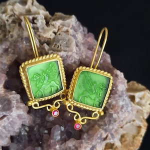 May include: Gold earrings with green glass squares featuring a winged lion design. The earrings have a dangling pink stone accent.