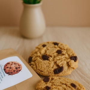 Peut inclure: Deux cookies ronds dorés aux pépites de chocolat noir visibles. Un cookie est entier et l'autre est cassé en deux. Un sachet en papier kraft avec une étiquette blanche illustrée d'un cookie est placé à proximité.