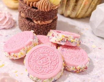 Może przedstawiać: Zbliżenie czekoladowego ciasta z lukrem i dodatkami oraz różowych ciasteczek Oreo. Ciasto ma warstwy czekoladowego ciasta z lukrem i jest zwieńczone preclem, maliną i innymi słodyczami. Różowe ciasteczka mają białe nadzienie i posypkę.