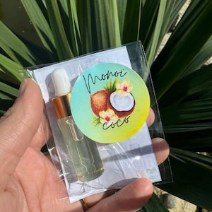 Può includere: Una piccola bottiglia di vetro con un contagocce, riempita con un liquido trasparente, è tenuta in una confezione di plastica trasparente. Un adesivo rotondo con una noce di cocco e fiori dice "Monoi coco".