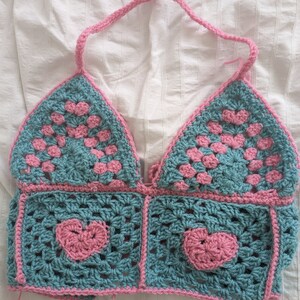 Könnte beinhalten: Ein gehäkeltes Halstuch mit einem rosa und türkisfarbenen Farbschema. Das Oberteil hat ein Herzdesign in der Mitte und eine Dreiecksform oben.