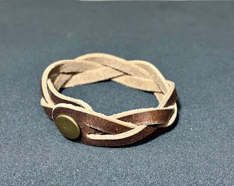 May include: Braided leather bracelet in shades of brown and tan. The bracelet features a woven design with a brass-colored button clasp. The accessory is a neutral color and suitable for everyday wear.