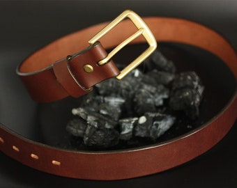 May include: A brown leather belt with a brass buckle. The belt is lying on a black surface with charcoal pieces.
