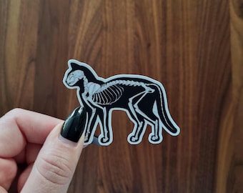 Könnte beinhalten: Schwarz-weißer Aufkleber eines Katzenskeletts. Die Katze steht auf allen vieren und blickt nach rechts. Der Aufkleber befindet sich auf einer Holzoberfläche.