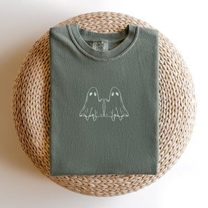 May include: A green t-shirt with a white outline of two ghosts holding hands. The t-shirt has the text "COMFORT COLORS" on the tag.