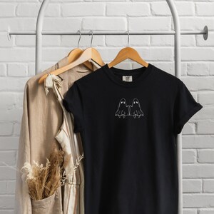 May include: Black t-shirt with a white outline of two ghosts holding hands. The shirt is hanging on a wooden hanger.