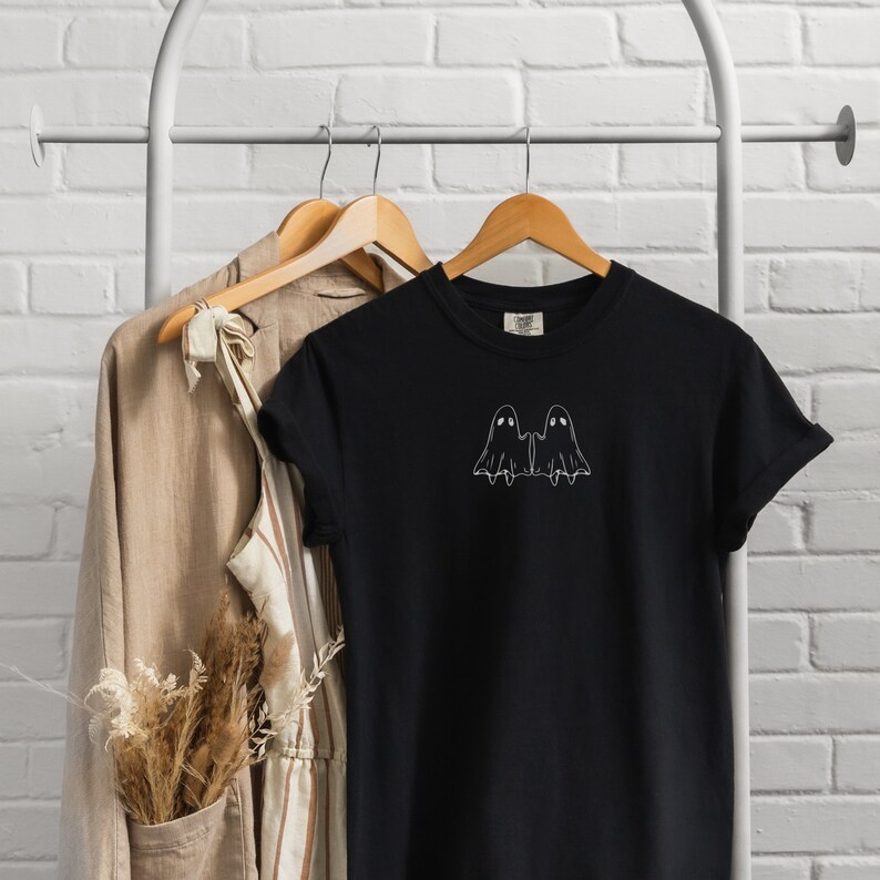 May include: Black t-shirt with a white outline of two ghosts holding hands. The shirt is hanging on a wooden hanger.