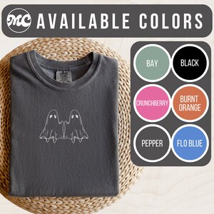 May include: A dark gray t-shirt with a white outline of two ghosts holding hands. The shirt is available in 10 colors: Bay, Black, Crunchberry, Burnt Orange, Pepper, Flo Blue, Heather Grey, Navy, Olive, and White.