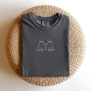 May include: A dark gray t-shirt with a white embroidered design of two ghosts holding hands. The shirt has a crew neck and is made of a soft, comfortable fabric.