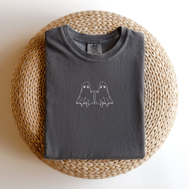 May include: A dark gray t-shirt with a white embroidered design of two ghosts holding hands. The shirt has a crew neck and is made of a soft, comfortable fabric.