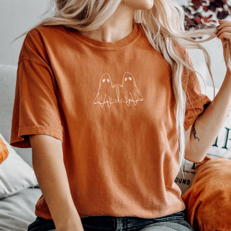 May include: A rust-colored t-shirt with a white outline of two ghosts holding hands. The ghosts have simple eyes and smiles.