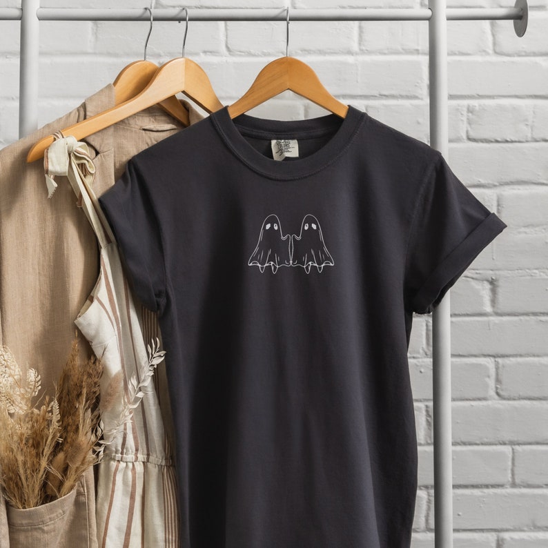 May include: A black t-shirt with a white outline of two ghosts holding hands. The ghosts have simple eyes and are facing each other.