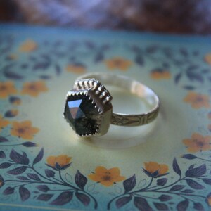 May include: A silver ring with a black gemstone set in a faceted, octagonal shape. The ring has a textured band with a decorative design.