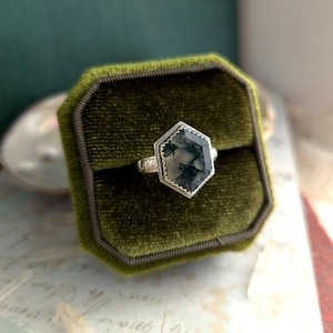 May include: A silver ring featuring a hexagonal moss agate gemstone, presented in a green velvet ring box. The moss agate displays green and white patterns. The ring's setting has a decorative bezel.