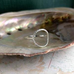 May include: A silver ring with a textured, open circle design. The ring is set against a pearlescent seashell, creating a contrast of textures and colors. The ring's band is thin and delicate.