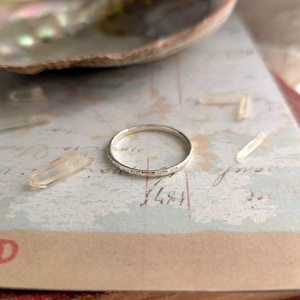 Puede incluir: Un anillo de plata con una banda texturizada descansa sobre un fondo de papel vintage. El anillo está junto a varios cristales de cuarzo transparente y una concha. El diseño del anillo es simple y elegante.