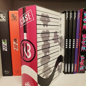 Puede incluir: Una caja rosa y negra con el texto "CASE 1/3" y una lupa. La caja está en un estante con otras cajas que tienen el texto "Stray Kids The 3rd Album" y "Stray Kids" en ellas.