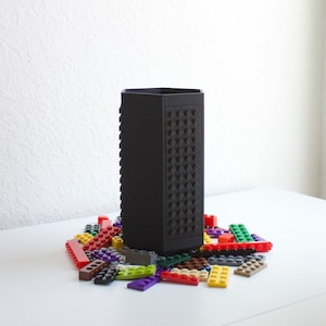 May include: A black, rectangular pencil holder with a Lego-like design, surrounded by colorful Lego bricks. The pencil holder has a textured surface with circular studs. The bricks are red, green, yellow, purple, and brown.