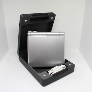 Op de afbeelding: Een zilveren handheld-spelconsole met een zwarte draagtas en een witte oplaadkabel.