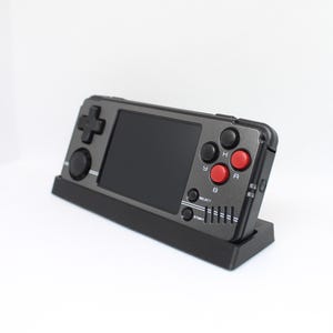 May include: A black and silver handheld video game console with red buttons. The console is on a black stand and has a screen, a directional pad, and buttons labelled "A", "B", "Select", and "Start".