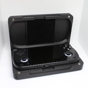Op de afbeelding: Een zwarte handheld gameconsole in een bijpassende zwarte beschermhoes. De console heeft een scherm, een navigatiepad en knoppen. De hoes is open en toont de console, die is vastgezet met kleine metalen sluitingen.