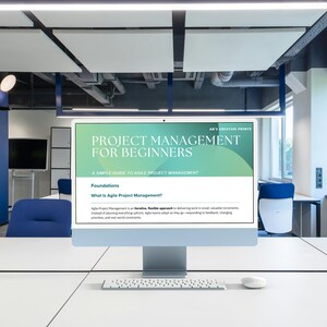 May include: A desktop computer displays a webpage titled "PROJECT MANAGEMENT FOR BEGINNERS." The screen shows text about Agile Project Management. A white keyboard and mouse sit on the white desk. The office has blue chairs and a blue wall.