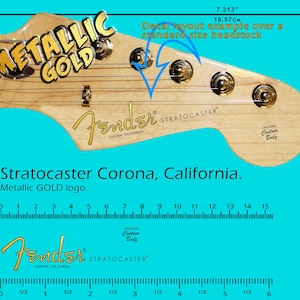 Puede incluir: Primer plano de una pala de guitarra con el logotipo Fender Stratocaster. La pala es de madera clara con herrajes dorados y las palabras "METALLIC GOLD". También se ve el texto "Stratocaster Corona, California".