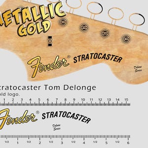 May include: A close-up of a guitar headstock with the words "METALLIC GOLD" in gold lettering. The Fender logo and "STRATOCASTER" are also visible. A ruler shows measurements in inches.