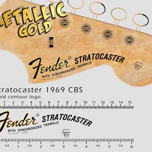 May include: A Fender Stratocaster guitar headstock with gold hardware and the words "METALLIC GOLD" in gold lettering. The text "Stratocaster 1969 CBS" is visible, along with a ruler in inches and centimeters.