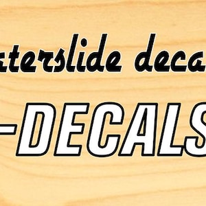 Może przedstawiać: Baner reklamowy dla Voodoo Decals, z tekstem "Premium quality waterslide decals!" i adresem strony internetowej. Logo Voodoo Decals zawiera postać szkieletu z kresk&oacute;wki. Tło to jasnobrązowe usłojenie drewna.