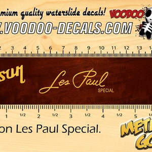 May include: A metallic gold Gibson Les Paul Special waterslide decal. The decal is on a dark brown background with the text "Gibson Les Paul Special" in gold script. The image also includes the text "All Gold."