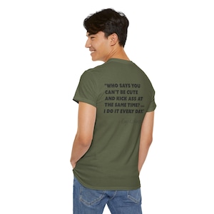 May include: Olive green t-shirt with black text on the back. The text reads: "WHO SAYS YOU CAN'T BE CUTE AND KICK ASS AT THE SAME TIME?... I DO IT EVERY DAY." The shirt is a short-sleeved crew neck.