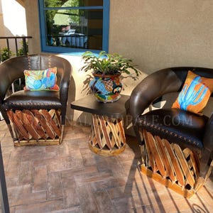May include: Two dark brown leather chairs with wooden bases and decorative pillows. A small table with a dark top and wooden base sits between the chairs. A potted plant sits on the table. The chairs are on a brick patio.