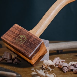 Puede incluir: Un mazo de madera con un mango marrón claro y una cabeza marrón oscuro. La cabeza está hecha de varias piezas de madera y tiene un logotipo dorado en la parte delantera. El mazo está sobre una superficie de madera con virutas de madera alrededor.